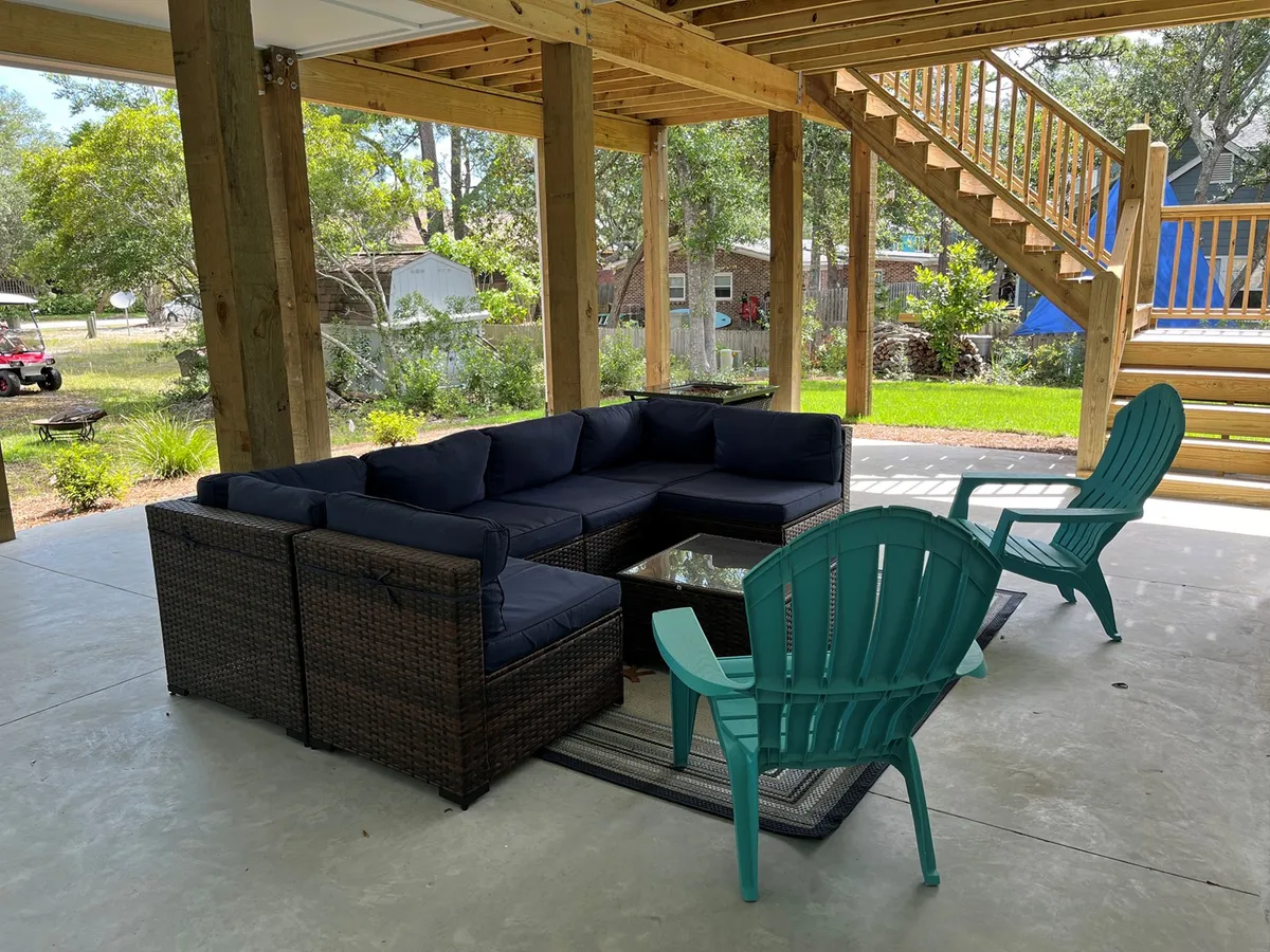 A shaded under-house patio area featuring a sectional sofa, glass coffee table, and turquoise Adirondack chairs, with a wooden staircase nearby. The space invites social gatherings, lounging, and afternoon snacks, shaded from the sun and set up for comfort and conversation.