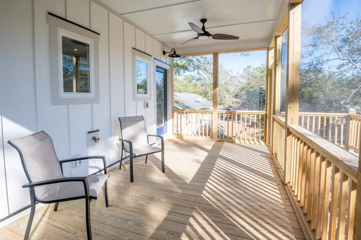 This image features a cozy screened porch with two inviting chairs, a ceiling fan overhead, and a blue door, with soft natural light streaming through the screens. The serene atmosphere suggests a perfect spot for enjoying a sunset cocktail, morning coffee, or board games while protected from insects and the elements.