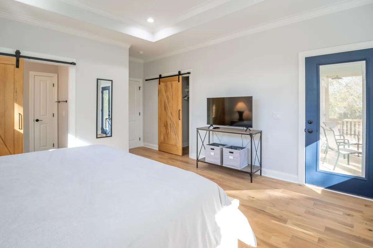 A tranquil guest bedroom featuring a queen-sized bed with crisp white bedding, a blue door leading out to a balcony, and a TV for added comfort. The space is bright, airy, and serene, offering a peaceful retreat for guests.