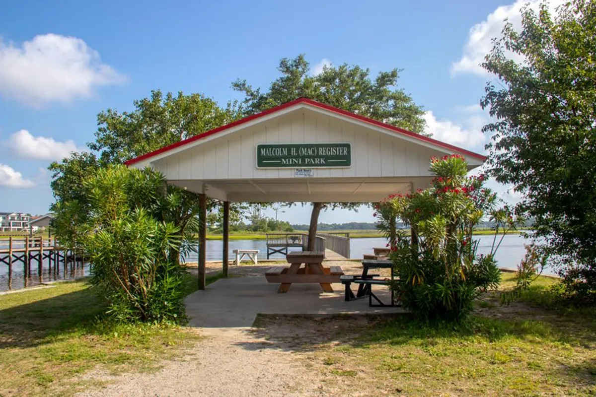 A scenic waterfront park with a pavilion and picnic tables surrounded by lush greenery. Families and groups can gather for picnics, play, and relaxation while enjoying peaceful views over the water. The setting suggests a space for community events and casual outdoor fun.