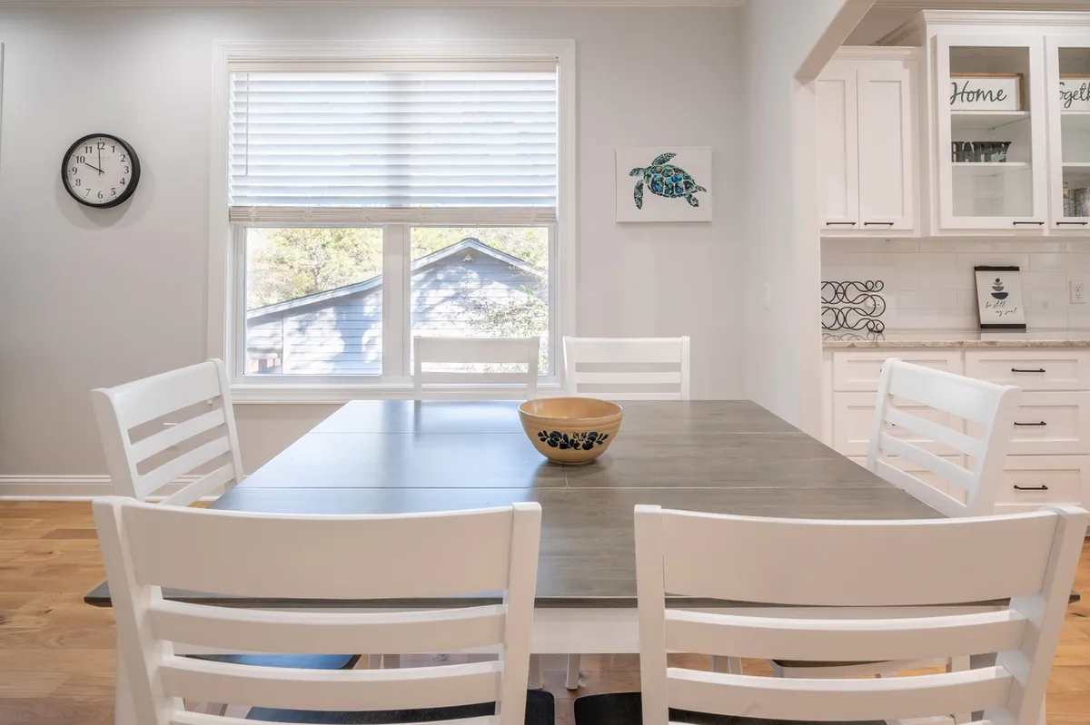 A welcoming dining area with a spacious table set under a window with a scenic view, centered by a decorative bowl. The space is well-lit and designed for family meals, gatherings, or game nights, with an inviting, homey atmosphere.
