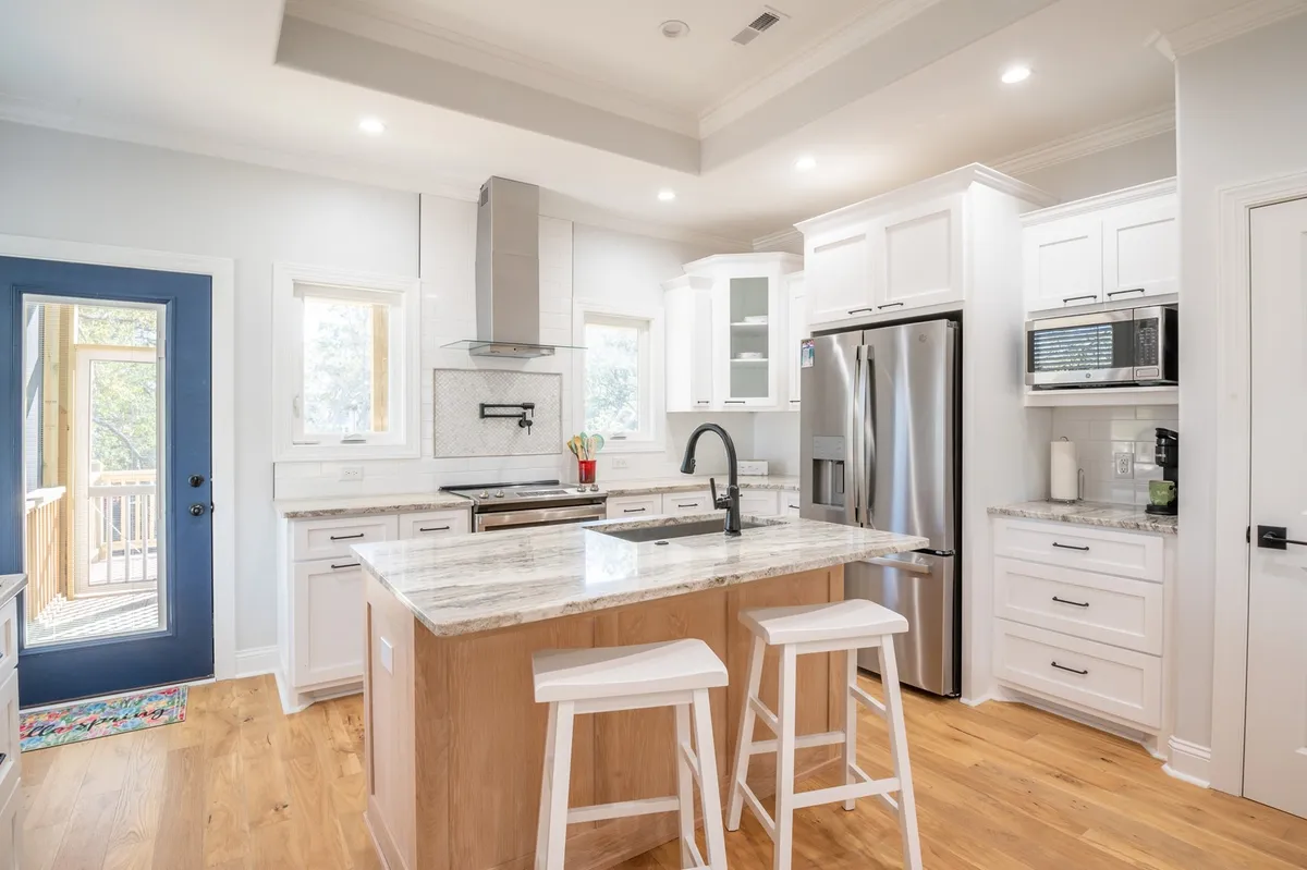A warm, open-concept kitchen featuring a central island with a granite countertop, surrounded by gleaming stainless steel appliances and a cheerful blue door. The natural light and open plan make it perfect for both quick breakfasts and festive gatherings.