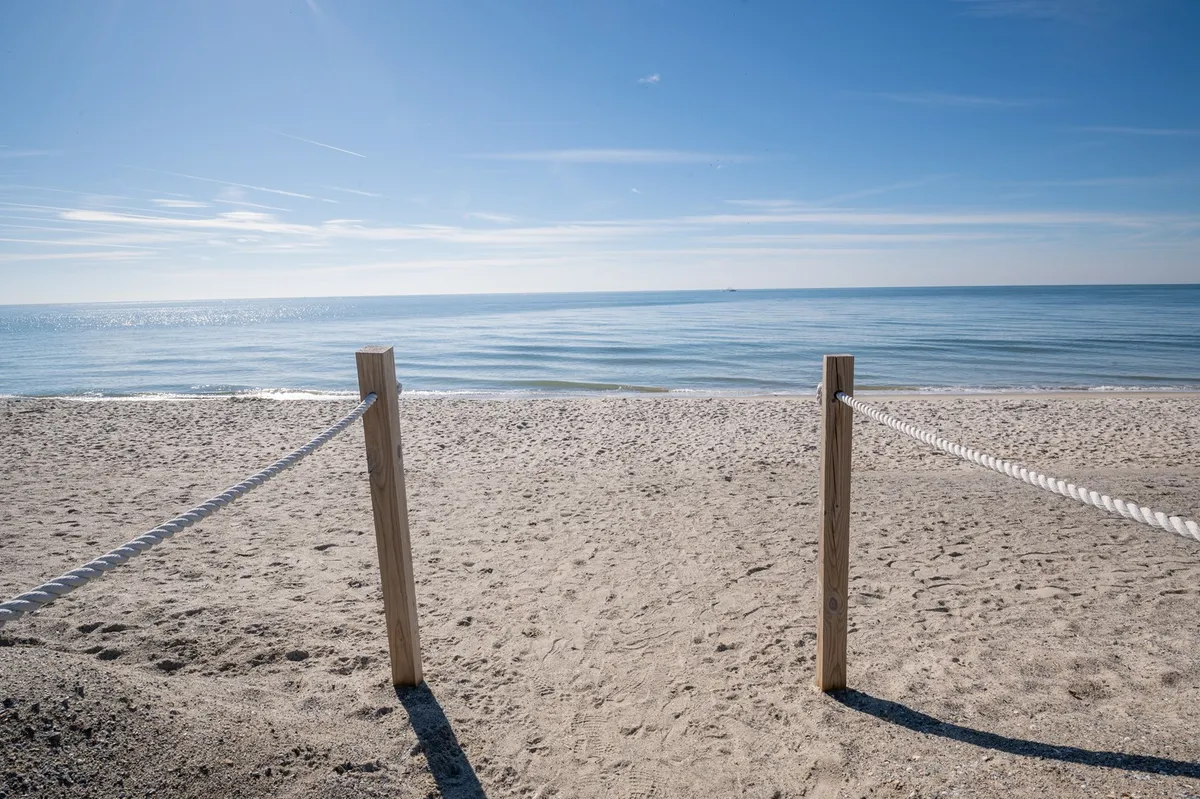 A tranquil sandy beach with a rope fence and calm ocean waves, bathed in soft sunlight. The wooden posts and gentle surf evoke a sense of peaceful isolation, perfect for leisurely walks, sunbathing, and beachcombing.