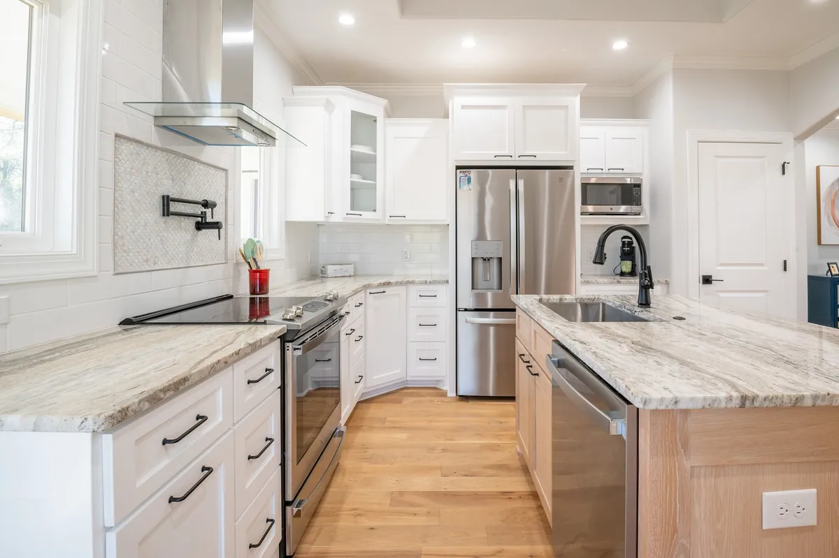 A stylish kitchen scene highlighting granite countertops and a large kitchen island, surrounded by stainless steel appliances and white custom cabinetry. The space feels open and welcoming, suitable for meal prep or entertaining guests.
