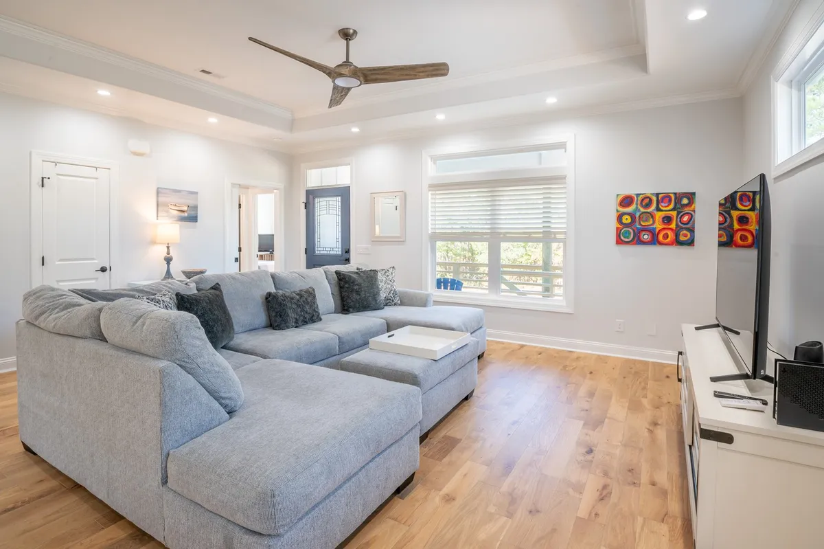A spacious and inviting great room featuring a large plush sectional sofa, TV unit, modern artwork on the walls, and a big window with blinds letting in soft natural light. The open plan allows ample room for gatherings, relaxation, and family activities, with stylish yet comfortable furnishings and accents.