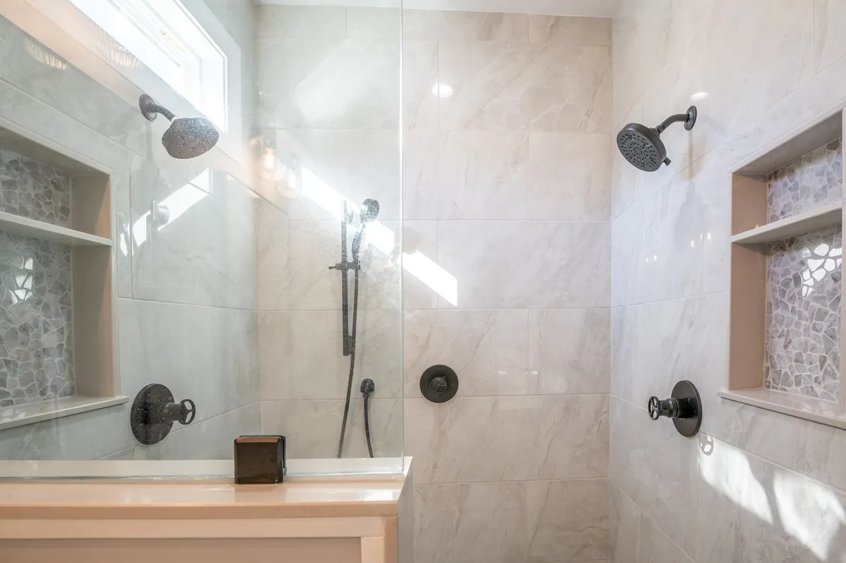 A modern ensuite bathroom with a glass-enclosed, zero-entry dual shower, textured tile walls, dual showerheads, and built-in shelving for added convenience. The overall design is spa-like and emphasizes luxury and accessibility.