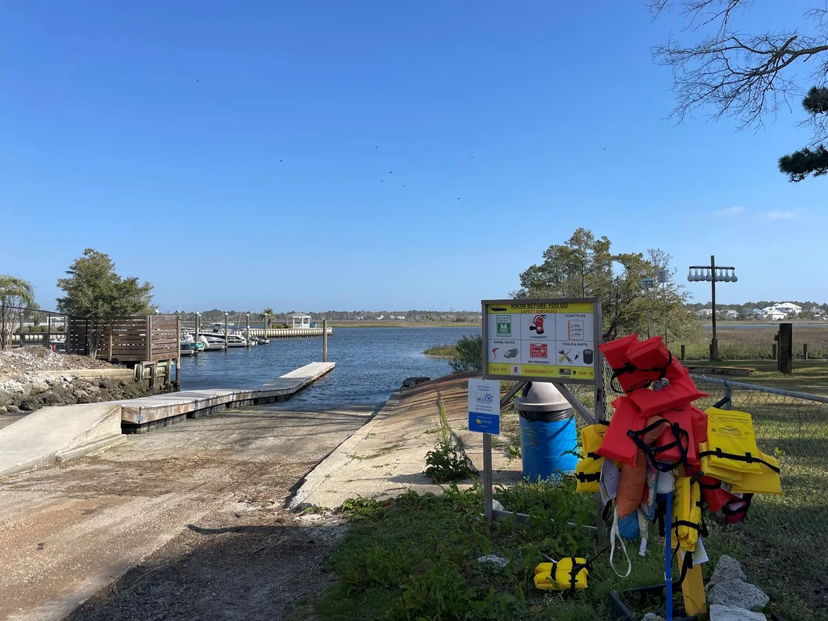 A lively marina scene showing a boat ramp, life jackets, informational signage, and a glimpse of the water and marina in the background. The area is set up for launching boats, kayaking, fishing, and waterfront recreation, with a welcoming, recreational vibe.