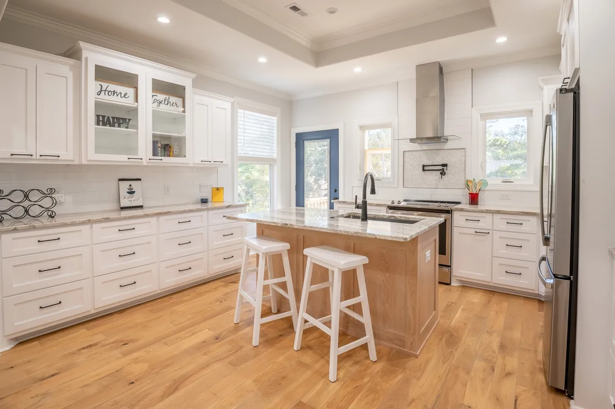A bright, inviting kitchen with a spacious island at its center, topped with sleek granite countertops. The kitchen features modern stainless steel appliances, white custom cabinetry, and a prominent blue door in the background. The space is well-lit and designed with gathering in mind, offering plenty of room for cooking, prepping, and socializing.
