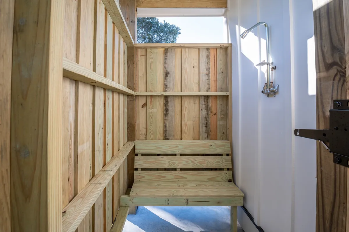 A serene outdoor shower area with wooden walls, a wooden bench, and a modern shower fixture, surrounded by natural textures. This spot offers a calming, nature-connected experience for rinsing off after the beach, emphasizing relaxation and practicality.