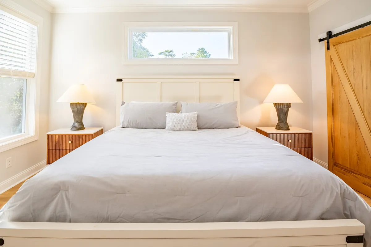 A serene bedroom featuring a king-sized bed with cozy bedding, soft lighting from matching lamps, a window allowing natural light to pour in, and a rustic barn door leading to an ensuite area. The spacious layout hints at comfort and privacy, ideal for a restful retreat.